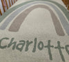 Close-up of a knitted blanket with a rainbow design and the name 'Charlotte' across the bottom. This blanket can easily be personalised with Babies name. The perfect gift for baby. Knitted in Oatmeal, Pale Grey and Fawn.