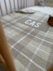 A beige and brown plaid patterned blanket with the name knitted on it, placed on a crib or cot. Available in Cot and Bassinet sizes.Knitted in Australia. The Original Personalised Blanket