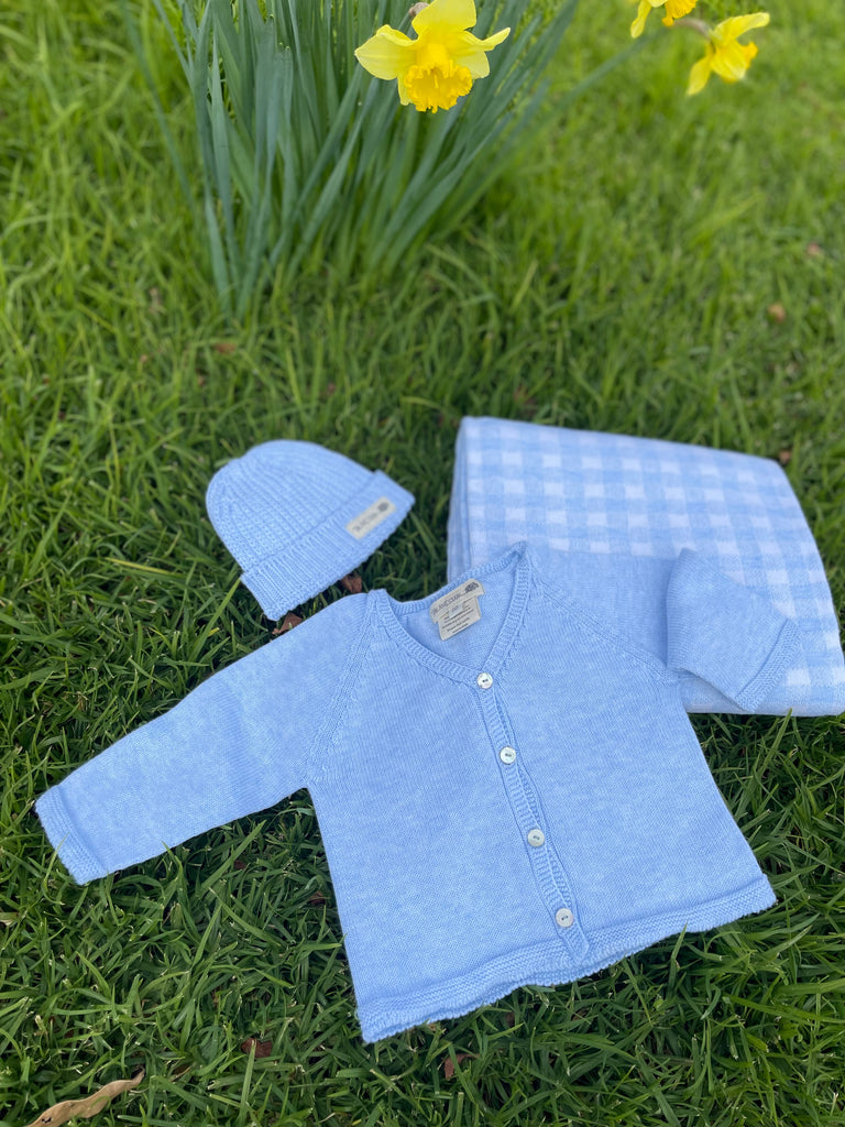 A blue fine knit cardigan, chunky knit beanie, and gingham cot blanket laid out on grass with daffodils in the background.
