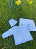 A blue fine knit cardigan, chunky knit beanie, and gingham cot blanket laid out on grass with daffodils in the background.