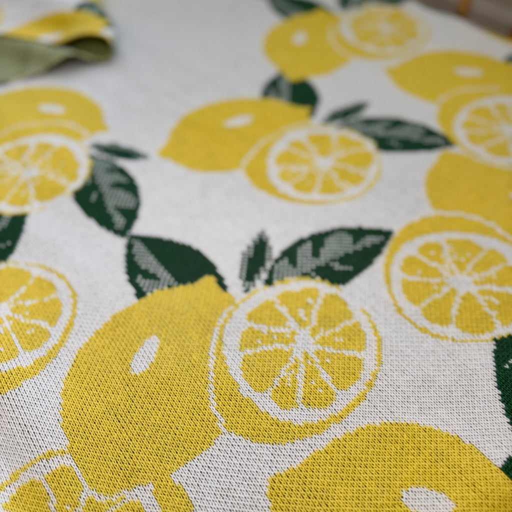Close-up of a knitted blanket with a citrus fruit pattern, predominantly yellow with green leaves. Organic Cotton