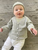 A baby wearing a light green, fine knit cardigan with natural shell buttons, paired with white pants, against a wooden backdrop.