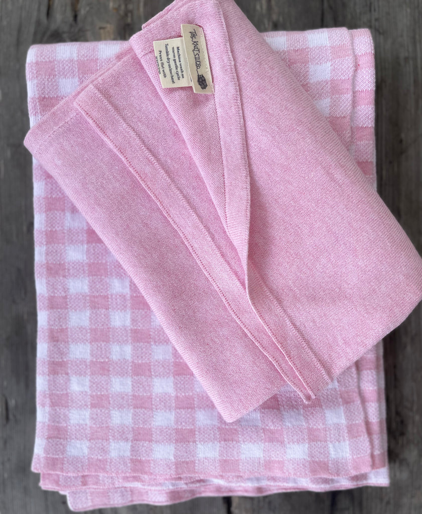 Folded pink and white gingham cot blanket and swaddle, placed on a wooden surface. Knitted in 100% Organic Cotton this giftset is as soft as it is sweet.