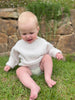A baby sitting on grass wearing a white ribbed merino knit pull-over jumper.